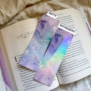 May include: Two decorative bookmarks with a marbled design in shades of purple, blue, and gold, resting on an open book. One bookmark is matte, the other is holographic. The text "MATTE" and "HOLOGRAPHIC" are visible.