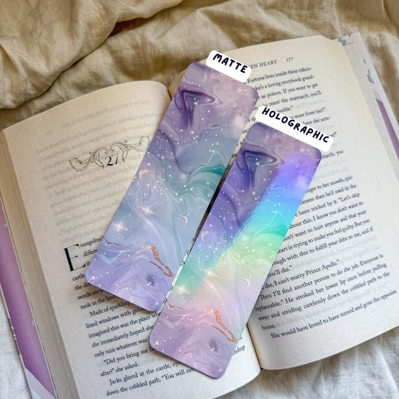 May include: Two decorative bookmarks with a marbled design in shades of purple, blue, and gold, resting on an open book. One bookmark is matte, the other is holographic. The text "MATTE" and "HOLOGRAPHIC" are visible.