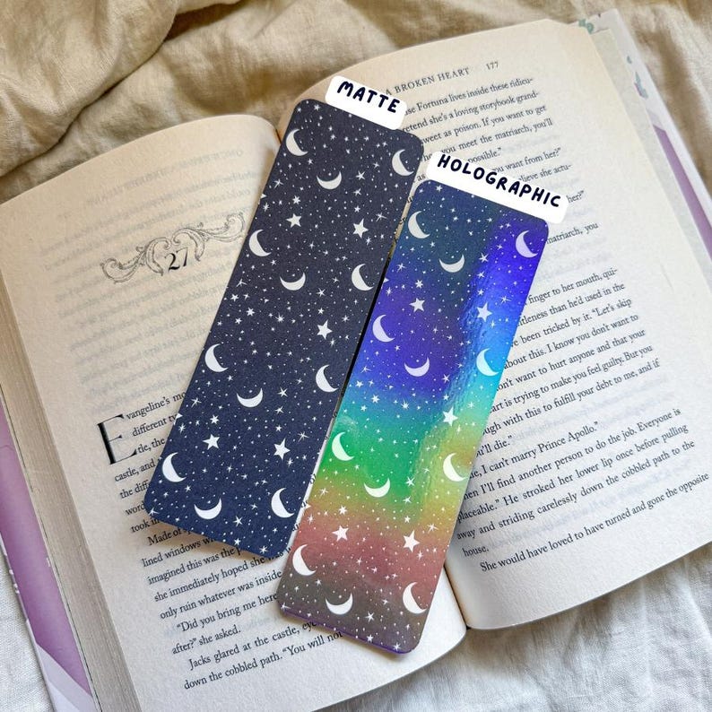 May include: Two decorative bookmarks with celestial designs. One is matte navy blue with white stars and crescent moons. The other is holographic with a rainbow gradient and the same celestial pattern. The bookmarks are placed on an open book.