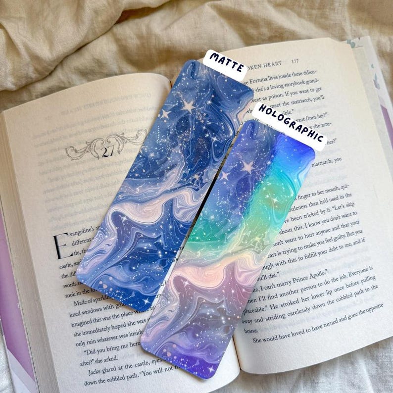 May include: Two rectangular bookmarks with celestial designs rest on an open book. One bookmark has a matte finish with a blue and white swirling pattern and stars. The other is holographic with a rainbow gradient and stars. Text on the bookmarks reads "MATTE" and "HOLOGRAPHIC".