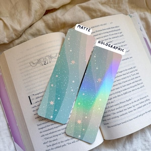 May include: Two rectangular bookmarks with a celestial design. One is matte with a light blue background and white stars. The other is holographic with a rainbow gradient and stars. The bookmarks are resting on an open book.