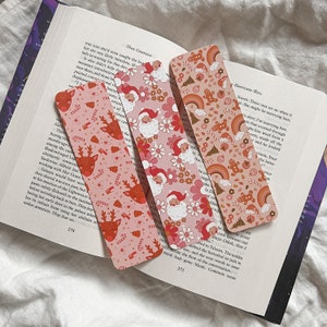May include: Three pink and orange Christmas-themed bookmarks with a repeating pattern of reindeer, Santa Claus, candy canes, and snowflakes. The bookmarks are made of metal and have a slightly textured surface.