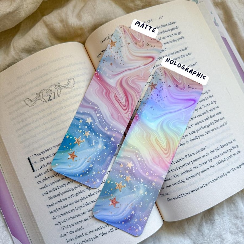 May include: Two decorative bookmarks with a swirling marble design in pastel colors, featuring gold star accents. One is matte, the other holographic. The bookmarks are placed on an open book.
