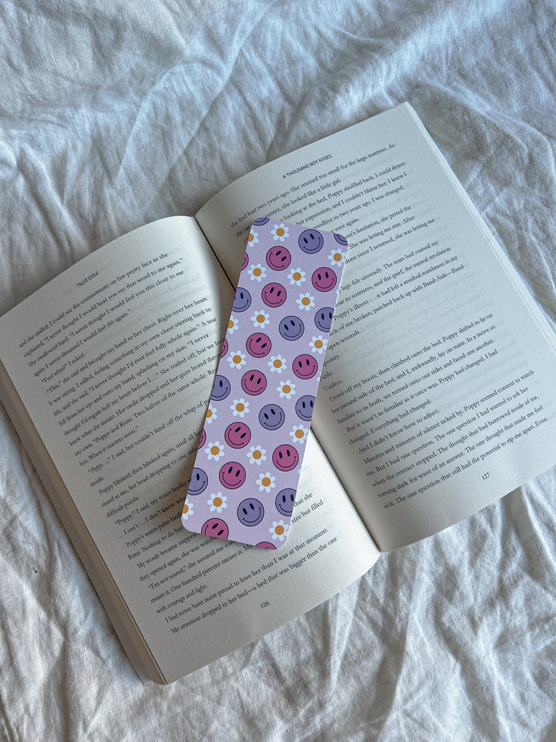May include: A purple and pink bookmark with a repeating pattern of white flowers and smiling faces. The bookmark is made of sturdy cardstock and is perfect for marking your place in a book.