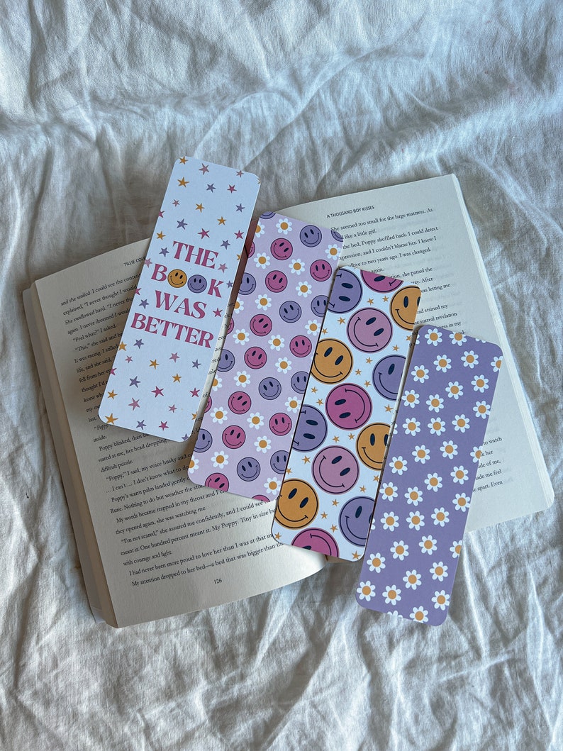 May include: Four colorful bookmarks with different designs. The first bookmark has a white background with pink and yellow stars and the text "The Book Was Better". The second bookmark has a purple background with white daisies and a pink smiley face. The third bookmark has a pink background with white daisies and a yellow smiley face. The fourth bookmark has a light purple background with white daisies and a pink and blue smiley face.