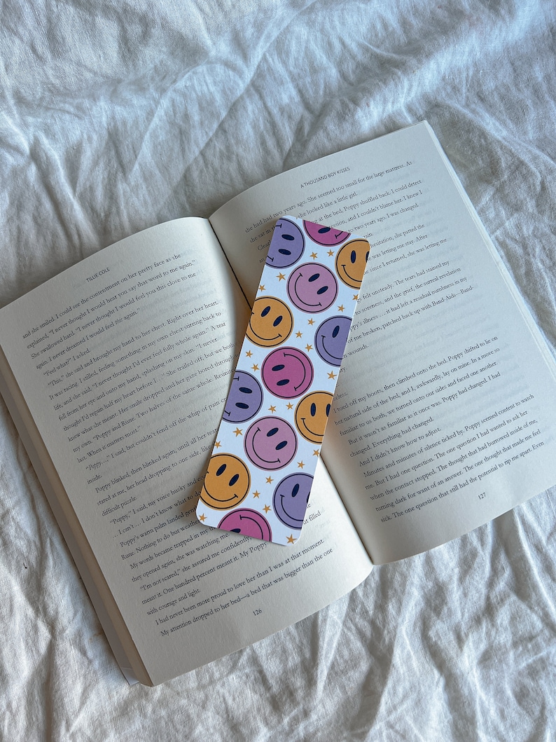 May include: A close-up of a book with a bookmark featuring a white background with a repeating pattern of colorful smiley faces and stars. The bookmark is placed on a page with text about a character named Poppy.