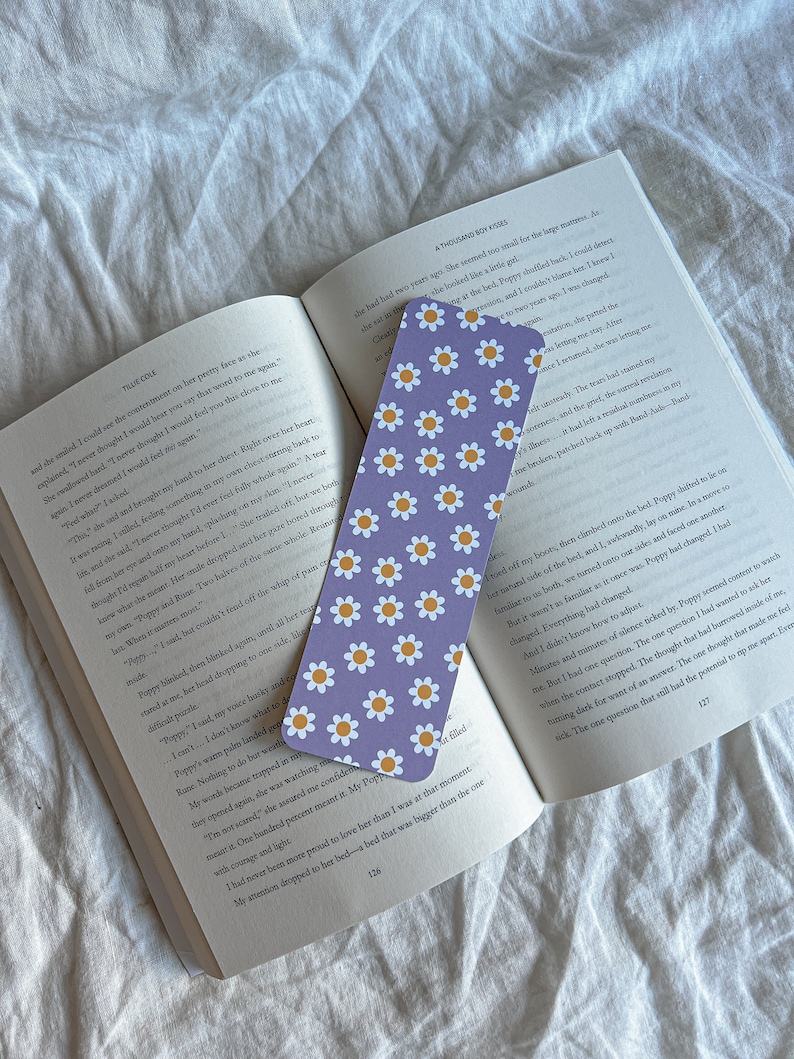 May include: A purple and white bookmark with a daisy pattern. The bookmark is on top of an open book with text about a character named Poppy.