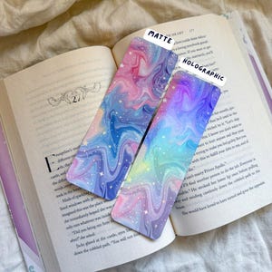 May include: Two colorful bookmarks with a swirling, galaxy-like design in shades of pink, blue, and purple, with white star accents. One is matte, the other holographic. The bookmarks are resting on an open book.