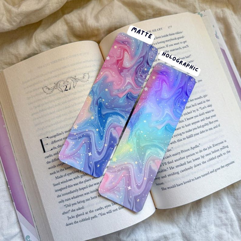 May include: Two colorful bookmarks with a swirling, galaxy-like design in shades of pink, blue, and purple, with white star accents. One is matte, the other holographic. The bookmarks are resting on an open book.