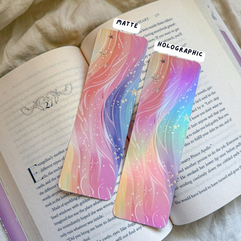 May include: Two colorful bookmarks with a rainbow design and star accents are displayed open on a book. One bookmark has a matte finish, and the other is holographic. The text "MATTE" and "HOLOGRAPHIC" are visible.