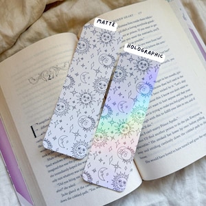 May include: Two celestial-themed bookmarks with sun, moon, and star patterns. One is matte, the other holographic. The bookmarks are placed on an open book, showcasing the product's use as a reading accessory.
