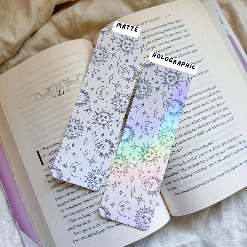 May include: Two celestial-themed bookmarks with sun, moon, and star patterns. One is matte, the other holographic. The bookmarks are placed on an open book, showcasing the product's use as a reading accessory.