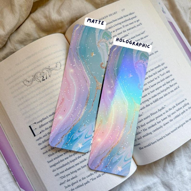 May include: Two decorative bookmarks with a pastel watercolor design. One is matte, the other holographic. Both have a swirling pattern in shades of blue, purple, and pink, with gold glitter accents and small star details.