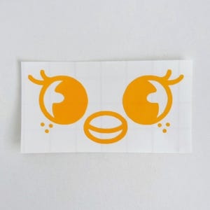 May include: A die-cut sticker featuring a stylized face with large, orange eyes, a small mouth, and three dots on each side. The design is in a bright orange color and is set against a white background.