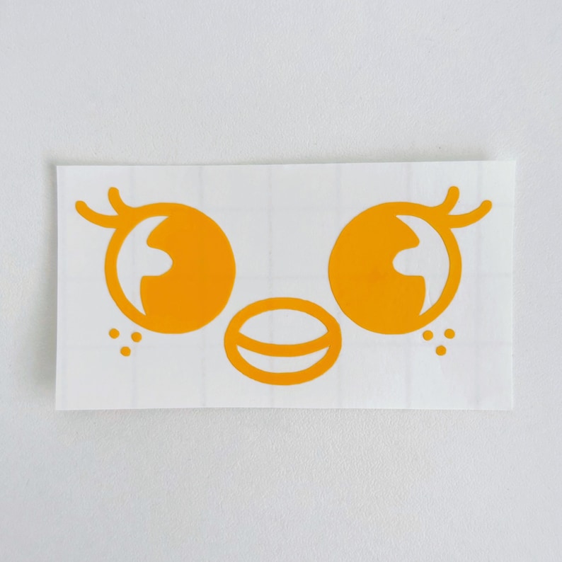 May include: A die-cut sticker featuring a stylized face with large, orange eyes, a small mouth, and three dots on each side. The design is in a bright orange color and is set against a white background.