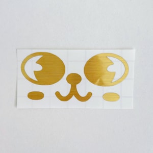 May include: Gold-colored sticker featuring a stylized bear face. The design includes two large oval eyes, a small nose, and two small cheek accents. The sticker is on a white background.