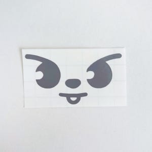 May include: A gray sticker featuring a cartoon face with angry eyes, a small nose, and a tongue sticking out. The sticker is rectangular and has a grid pattern on the backing.