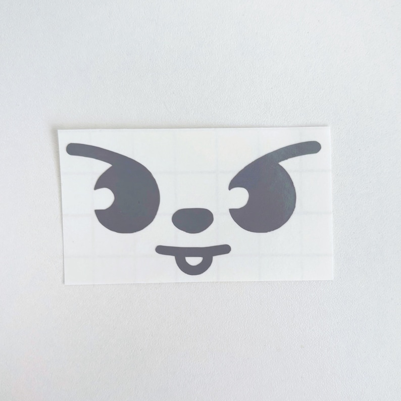 May include: A gray sticker featuring a cartoon face with angry eyes, a small nose, and a tongue sticking out. The sticker is rectangular and has a grid pattern on the backing.