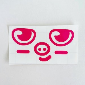 May include: A sticker featuring a cartoon pig face with bright pink eyes, a nose with two dots, a curved mouth, and two horizontal lines. The pig face is on a white background.