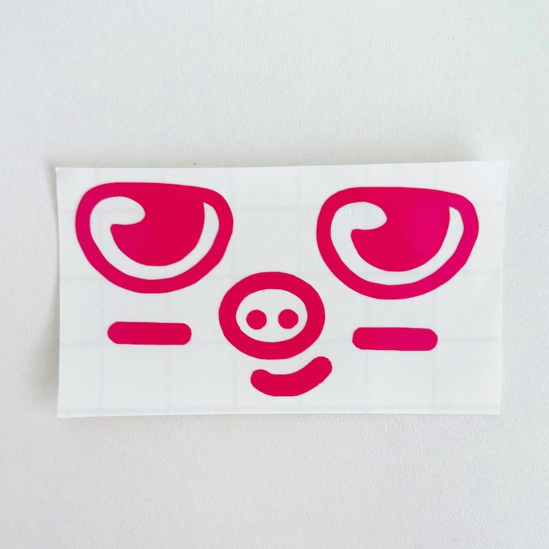 May include: A sticker featuring a cartoon pig face with bright pink eyes, a nose with two dots, a curved mouth, and two horizontal lines. The pig face is on a white background.
