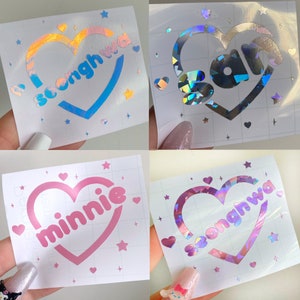 May include: Four different holographic vinyl stickers with heart-shaped designs. Each sticker features a different name: "Seonghwa", "San", "Minnie", and "Seonghwa". The stickers are on a white background with small stars and hearts.