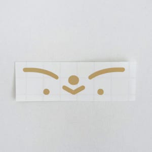 May include: A rectangular sticker with a gold-colored minimalist face design. The design includes two curved lines for eyebrows, a circle for the nose, a downward-pointing chevron for the mouth, and two small circles for the eyes. The sticker is on a white background.