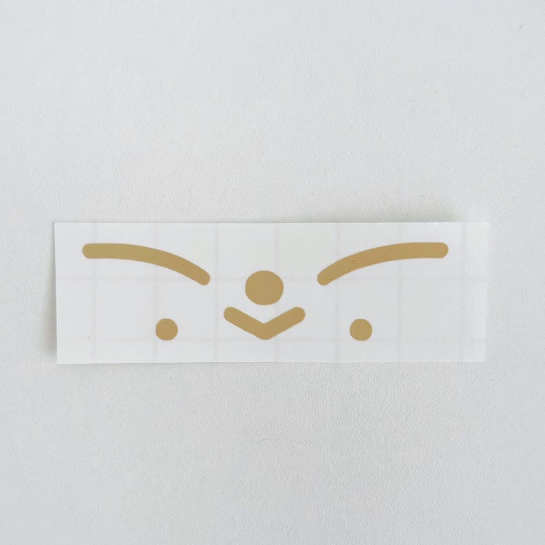May include: A rectangular sticker with a gold-colored minimalist face design. The design includes two curved lines for eyebrows, a circle for the nose, a downward-pointing chevron for the mouth, and two small circles for the eyes. The sticker is on a white background.