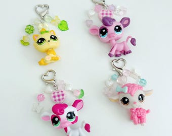 May include: Four keychains featuring stylised animal figures: a yellow cat, a pink cow, a white and pink cow, and a pink lamb. Each figure is attached to a silver heart-shaped clasp and decorated with various translucent beads, charms, and small plastic shapes.