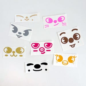 May include: A collection of colorful face stickers with various expressions. The stickers feature different eye shapes, noses, and mouths in shades of pink, gold, brown, gray, and black. Each sticker is rectangular and has a white background.