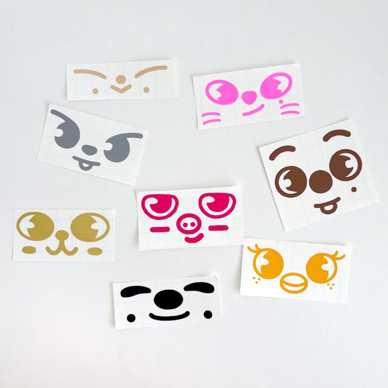 May include: A collection of colorful face stickers with various expressions. The stickers feature different eye shapes, noses, and mouths in shades of pink, gold, brown, gray, and black. Each sticker is rectangular and has a white background.