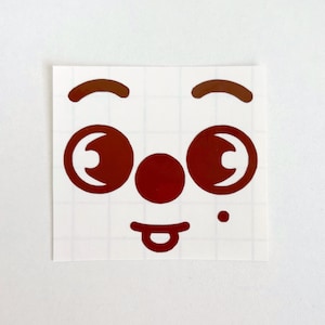May include: A square sticker featuring a cartoon face with brown eyes, eyebrows, nose, and a tongue sticking out. The sticker has a white background with a grid pattern.