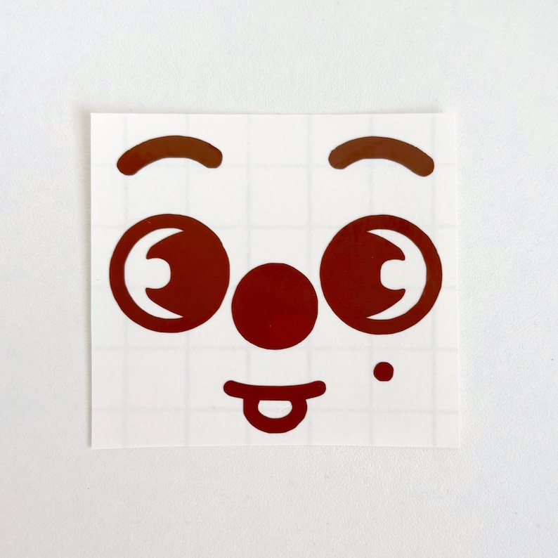 May include: A square sticker featuring a cartoon face with brown eyes, eyebrows, nose, and a tongue sticking out. The sticker has a white background with a grid pattern.