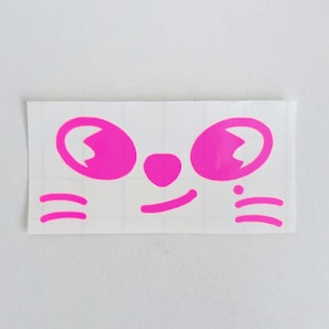 May include: A rectangular sticker featuring a cartoon face with bright pink details. The design includes large, oval eyes, a small nose, a smiling mouth, and whiskers. The sticker is on a white background.