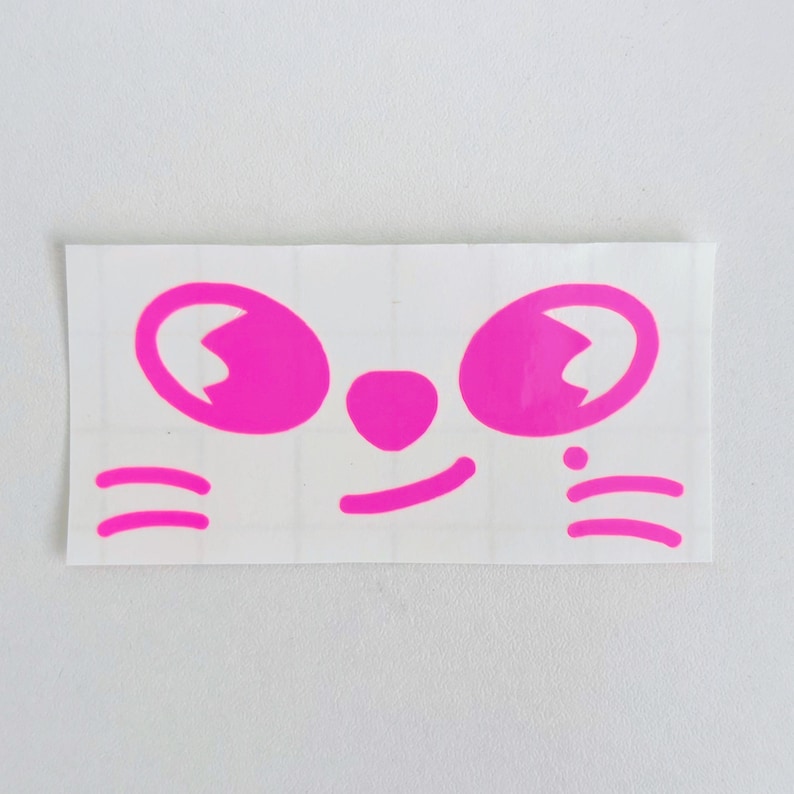 May include: A rectangular sticker featuring a cartoon face with bright pink details. The design includes large, oval eyes, a small nose, a smiling mouth, and whiskers. The sticker is on a white background.