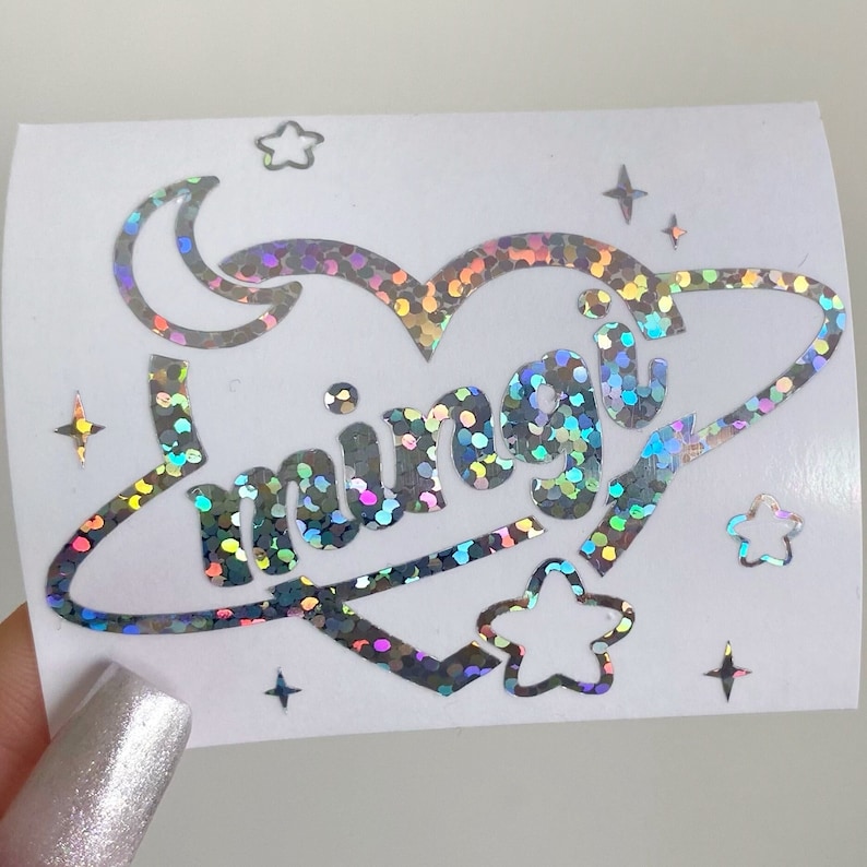 May include: A holographic sticker with a heart-shaped design, a crescent moon, and the word "miingiu" inside a planet ring. The sticker is surrounded by small stars.