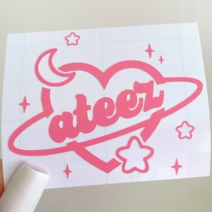 May include: Pink vinyl sticker with a heart-shaped planet design. The planet has the word "ateez" written on it in a retro font. The sticker also features a crescent moon and stars.