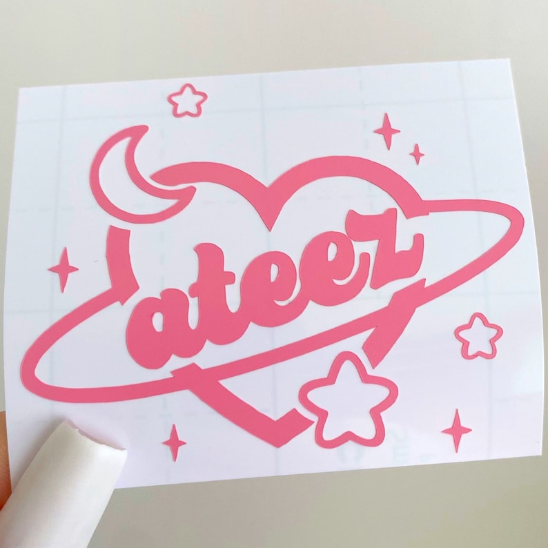 May include: Pink vinyl sticker with a heart-shaped planet design. The planet has the word "ateez" written on it in a retro font. The sticker also features a crescent moon and stars.
