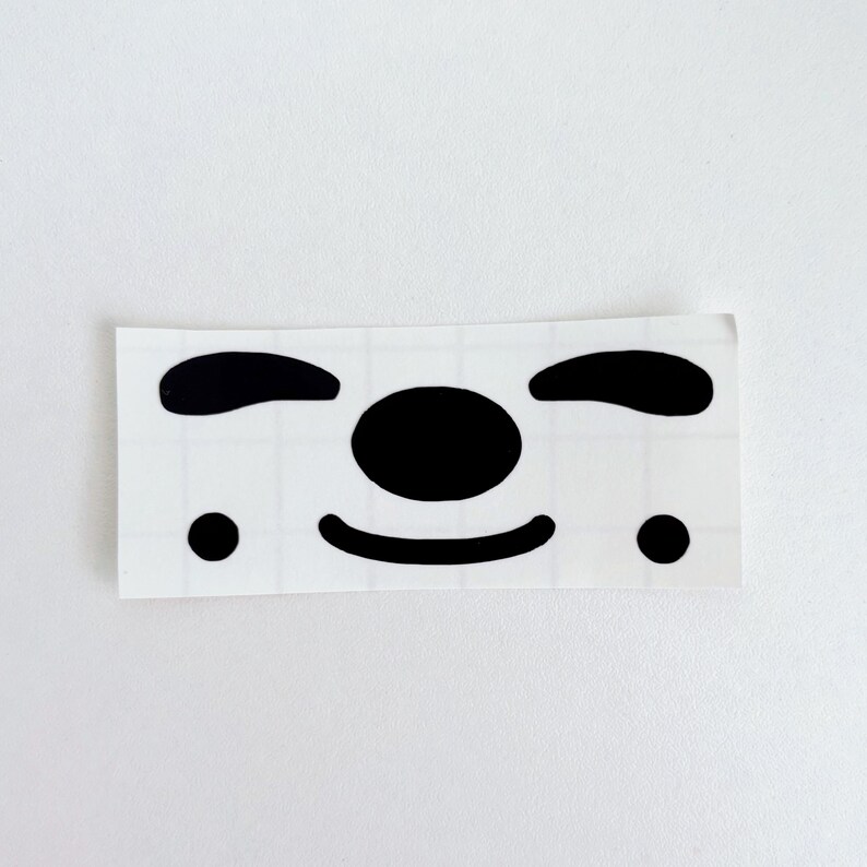 May include: A rectangular sticker featuring a minimalist face design. The face is composed of black shapes: two curved lines for eyebrows, a large circle for the nose, a curved line for the mouth, and two small dots for cheeks.