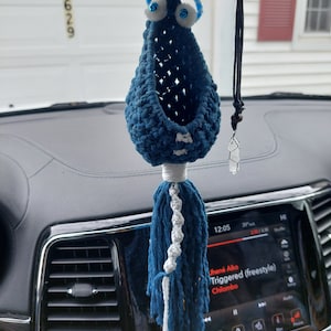 Może przedstawiać: Ręcznie robiony, niebieski urok do samochodu z białymi akcentami i ruchomymi oczami. Urok ma frędzel i wisi na lusterku wstecznym samochodu. W pobliżu wisi przezroczysty kryształowy wisiorek.