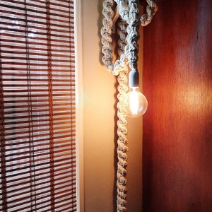 May include: A white rope light fixture with a light bulb turned on. The rope is knotted and wrapped around a wooden base. The light fixture is mounted on a wall.