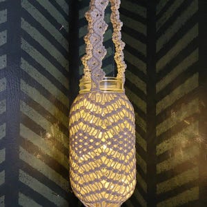 May include: A white macrame hanging lampshade with a glass jar in the center. The lampshade is hanging from a ceiling and is lit from inside, casting a warm glow.