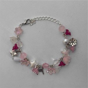 Op de afbeelding: Een zilveren bedelarmband met roze en witte bloem- en sterbedels, roze kralen en een kleine varkensbedel. De armband heeft een karabijnsluiting.