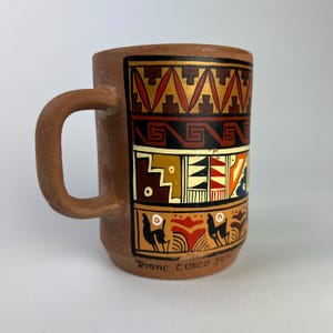 May include: A brown ceramic mug with a handle, featuring colorful geometric patterns in red, gold, black, and white. The mug has the text "Pisac Cusco Peru" at the bottom, and is likely handmade.