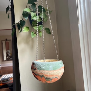 May include: A round, bowl-shaped hanging planter featuring a marbled pattern of sage green, cream, and burnt orange colors with a textured surface. The planter is suspended by three thin silver-toned metal chains attached to the rim.