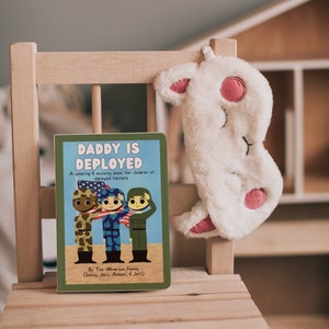 Op de afbeelding: Een groen boek met de titel "Daddy is Deployed" met een kleur- en activiteitenboek voor kinderen van ingezet personeel. Het boek bevat een illustratie van drie cartoon soldaten die salueren.