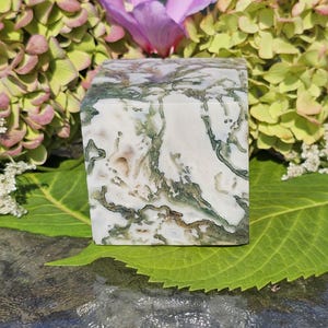May include: A white and green cube-shaped polished stone with a marbled pattern. The stone is sitting on a green leaf.