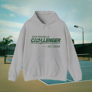 Könnte beinhalten: Ein grauer Kapuzenpullover mit dem Text "NEW ROCHELLE CHALLENGER" in Grün. Der Text befindet sich über einer grünen Linie mit dem Text "PRESENTED BY PHIL'S TIRETOWN" in kleinerer Schrift darunter.