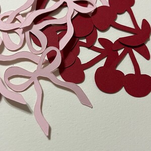 Puede incluir: Un mont&oacute;n de recortes de papel rosa y burdeos de cerezas y lazos. Los recortes est&aacute;n esparcidos sobre una superficie blanca.