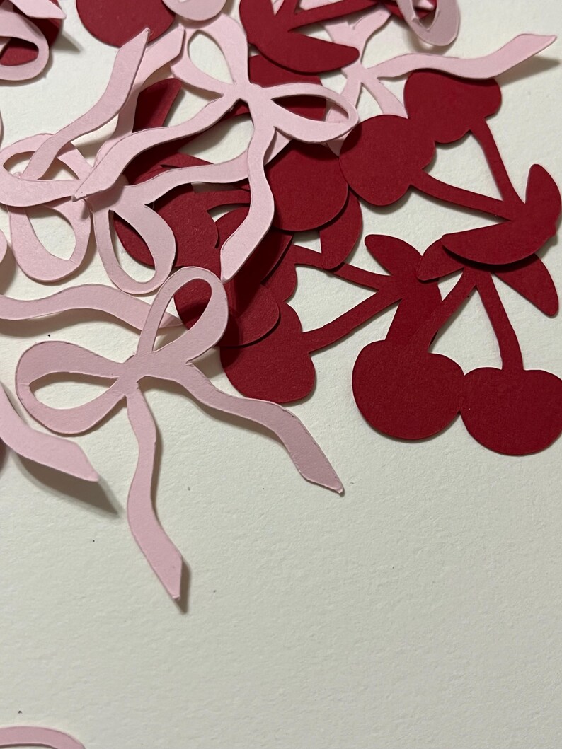 Puede incluir: Un mont&oacute;n de recortes de papel rosa y burdeos de cerezas y lazos. Los recortes est&aacute;n esparcidos sobre una superficie blanca.