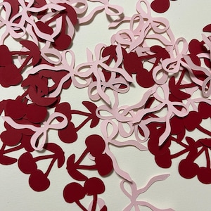 Puede incluir: Un mont&oacute;n de recortes de papel rojo y rosa de cerezas y lazos. Las cerezas son de color rojo oscuro y los lazos son de color rosa claro. Los recortes est&aacute;n esparcidos sobre un fondo blanco.
